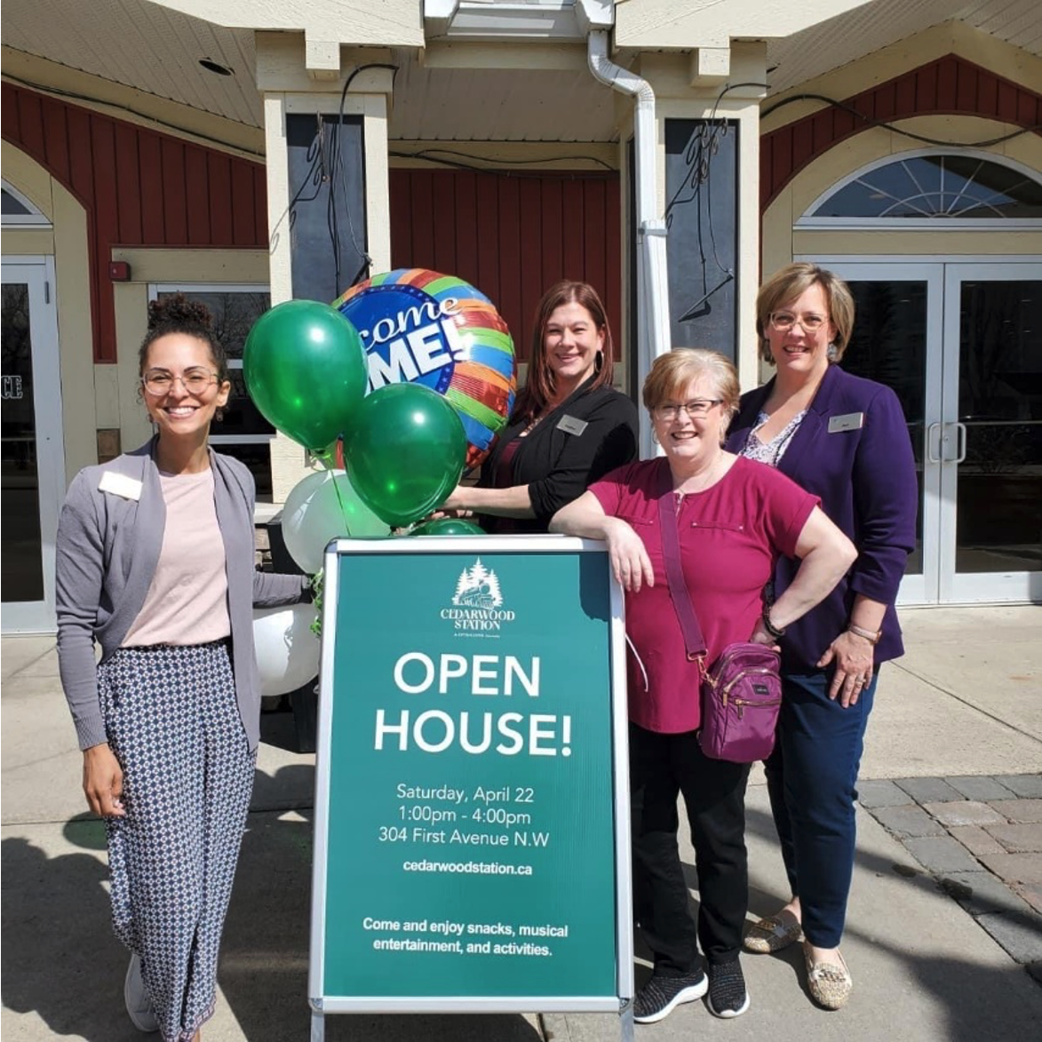 Four women stand outside of retirement community Cedarwood Station with balloons a sign advertising their open house.